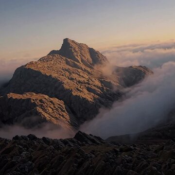 Low Angle of towering mountain peak kissed by dawn light, surrounded by misty clouds, with warm, golden color grading, soft sunlight breaking through, evoking resilience and triumph.

