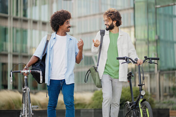 Two male colleagues walking together in the city pushing bicycles going to the office, talking and smiling