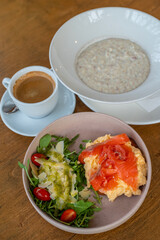 Delicious healthy breakfast spread featuring creamy oat porridge, scrambled eggs topped with smoked salmon, a fresh arugula salad with cherry tomatoes and parmesan, paired with a smooth café crema. 