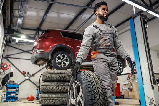 Young interracial car mechanic with tire walking at automotive car service and rolling it.