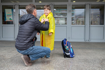 Father helps son put on raincoat after school. Parental care, rainy day.