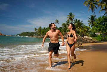 Happy couple runs on tropical beach, hand in hand. Healthy young man, woman in swimwear jog on...
