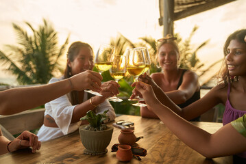 Friends toast wine glasses at sunny outdoor restaurant. Diverse women celebrate bachelorette party trip. Relaxed vibes and fun chat. Stylish setting with palm tree view.