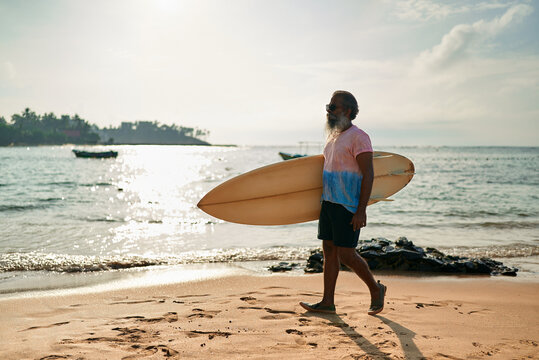Active senior ethnic man walks on sunny beach with surfboard. Elderly surfer enjoys ocean waves in vibrant lifestyle. Retirement adventure with surf culture. Promoting healthy aging in coastal scene.