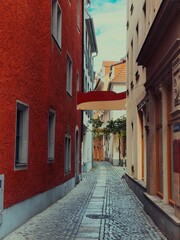 A charming narrow alley with cobblestone pavement, framed by colorful historic buildings. The red facade contrasts beautifully with the muted tones, creating a picturesque European street scene.