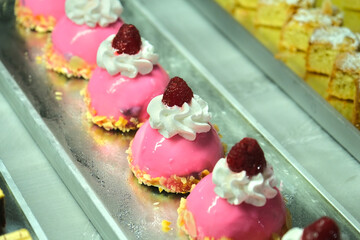 Delicious pink dome-shaped pastries with glossy glaze, whipped cream, a raspberry topping, and almond flakes on a tray in a bakery display.