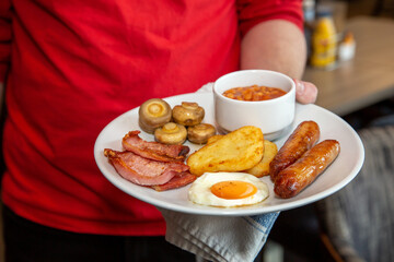 Full Irish breakfast served by waiter at a hotel with sausge, eggs, baked beans, bacon, hash browns and mushrooms.