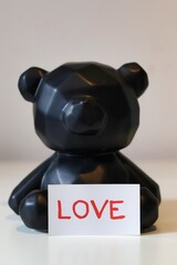 Minimalist figurine of a black teddy bear holding a card with the handwritten word 'love'