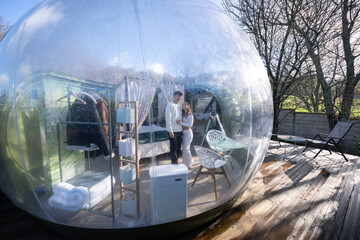 Romantic couple hugging inside transparent dome accommodation, featuring minimalist interior design and wooden terrace, experiencing intimate secluded lodging