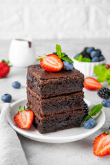 A stack of chocolate brownies with chocolate glaze, fresh berries and mint leaves on top on a white plate on a gray concrete background. Delicious dessert.