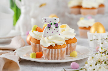 Easter cupcakes are decorated with cream, sugar bunnies, chocolate eggs, jelly carrots on a white plate. Easter food composition. Copy space.