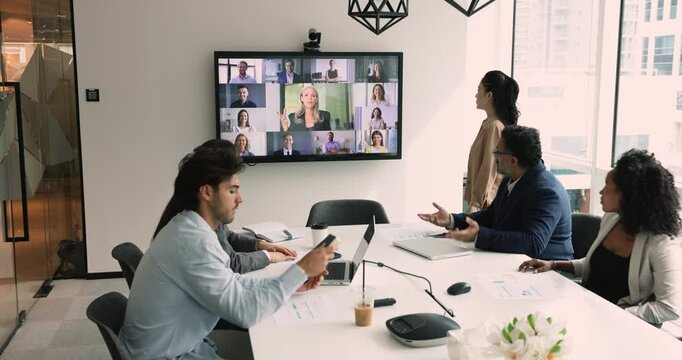 Diverse business team watching webinar of senior professional coach on large wall electronic board, having online video conference with remote colleagues, sitting at office meeting table