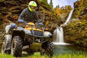 Man on ATV near waterfall. Guy is sitting on motorcycle SUV. Overcoming mountain river on quad bike. ATV driver in yellow helmet. Man on dirty ATV near mountains. Traveling on motorcycle SUVs