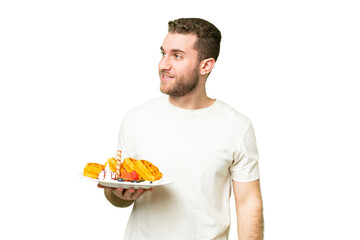 Young handsome blonde man holding waffles over isolated chroma key background looking to the side and smiling