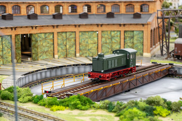 vintage green locomotive on a railway turntable in front of a historic brick depot with open green doors and lush greenery