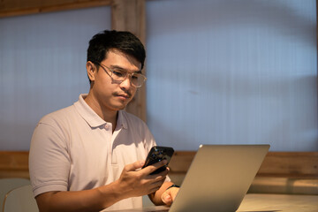 A man is sitting at a table with a laptop and a cell phone. He is looking at the cell phone while using the laptop
