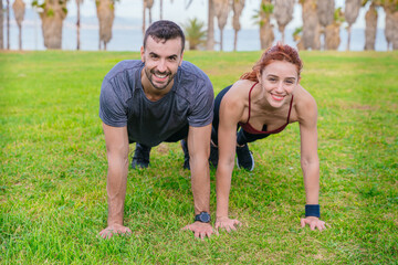 Smiling young man and woman doing push-ups together in a green park near the coast. Both are wearing sportswear and look motivated and energetic. The background features palm trees and nature.
