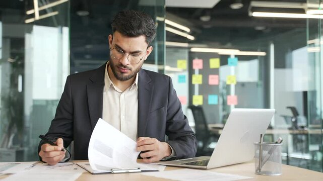 Businessman in a formal suit signing documents sitting at desk at workplace in a business office. Boss in glasses looks through the folder with documentation, checks and signs the contract with a pen