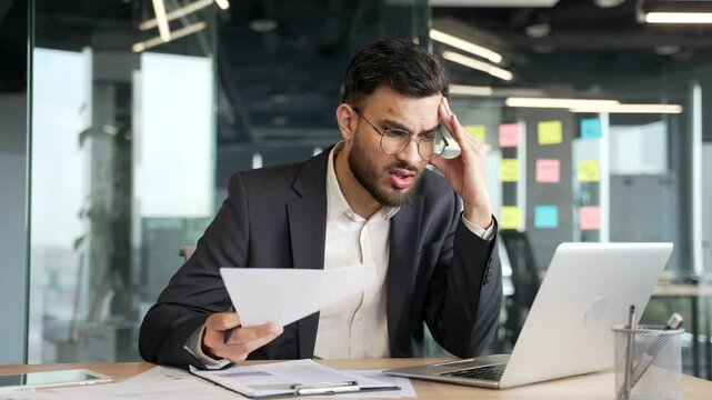 Frustrated businessman unhappy with financial results sitting at desk at workplace in business office. Upset puzzled financial investor in a formal suit unhappily reading document with bad indicators
