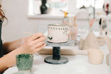 Artist is decorating and painting handmade ceramic clay cup in the creative workshop. Painter is drawing and adding unique designs to the earthenware.
