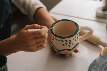 Potter draws an abstract design into earthenware cup. Experienced artist painting designs on ceramic cup in pottery workshop.