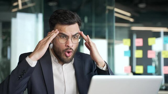 Upset worried businessman in a formal suit reads bad news on laptop sitting at workplace in business office. Frustrated entrepreneur is sad while reviewing negative information on a computer. Close up