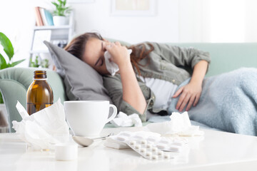 A young woman with the flu lying on the couch blows her nose with a tissue and holds her stomach in...