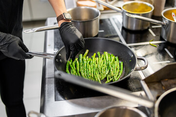 A chef wearing black gloves skillfully stirs green beans in a frying pan on a modern kitchen stovetop, surrounded by stainless steel cookware and vibrant ingredients.