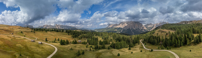 Expansive aerial view of the Pralongia Plateau in the Dolomites, Italy. Captures vibrant pastures, forested slopes, and the rugged Sella massif
