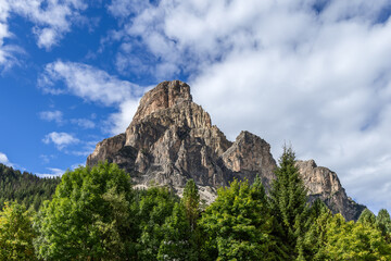 Stunning view of the Sassongher mountain, part of the Dolomites, surrounded by dense pine and deciduous trees, with dramatic cliffs and clouds