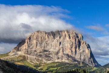 The towering Sassolungo in the Dolomites, Italy, dominates the landscape with steep rocky slopes and lush alpine terrain below a cloudy blue sky