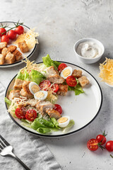 Caesar salad with fried chicken, cherry tomatoes, croutons, cheese chips, parmesan, lettuce and eggs in white plate on light background. Stylish restaurant serving