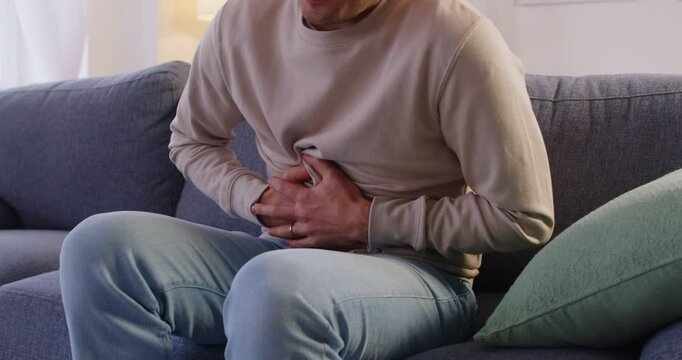 Cropped view of sick man feeling intense pain in his stomach, clutching his abdomen in discomfort. Unhealthy male person sitting on sofa at home, suffering from abdominal ache and strong cramps.
