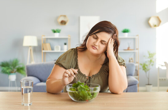 Overweight woman feeling frustrated while eating fresh vegetable salad. Unhappy fat female sitting at table with bowl of greens and glass of water, showing dissatisfaction with diet and healthy meal. - Powered by Adobe