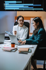 Two asian businesswomen are working together, discussing charts and using a laptop in a brightly lit office meeting room
