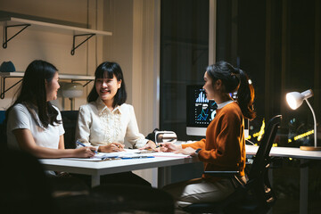 Fototapeta premium Three asian businesswomen working late in a brightly lit office, discussing charts on a computer screen and taking notes