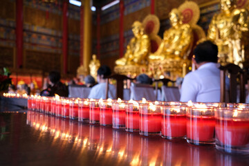 Rows of red candles flicker in front of golden Buddha statues inside a serene temple, as worshippers engage in prayer and meditation, creating a peaceful and spiritual atmosphere.