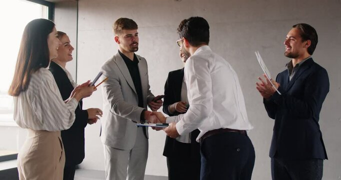 Business leader, manager or CEO acknowledging and praising employee for achieving excellent work results, shaking hands with him. Team of colleagues congratulating their colleague and applauding.