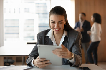 Positive successful young business woman watching online content on digital tablet, reading good news, enjoying Internet startup success, using digital gadget in presentation room, smiling