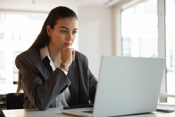 Focused pensive young businesswoman working at laptop in office, touching chin, thinking on online...