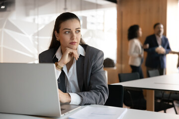 Serious dreamy young manager woman working at laptop in office co-working room, touching chin, looking away, thinking on solutions for teamwork, colleagues talking in blurred background