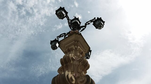 Upward view of Fuente de la Farola, highlighting its ornate stone base and historic four-arm lamp, framed by a cloudy bright sky.