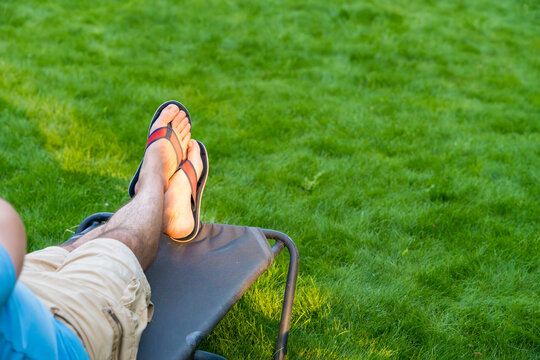 A man relaxing on a lounge chair with his legs crossed, wearing sandals, and enjoying summer sun