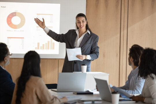 Confident young speaker woman presenting sales growth report to business team of colleagues, partners, investors, pointing hand at slide on white board, smiling, giving marketing training