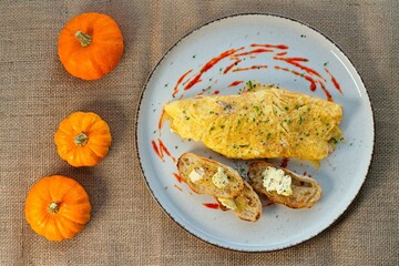 The omelette is presented on a rustic plate alongside grilled sourdough bread, butter, and chives, all set against a rustic tablecloth featuring pumpkin decorations.