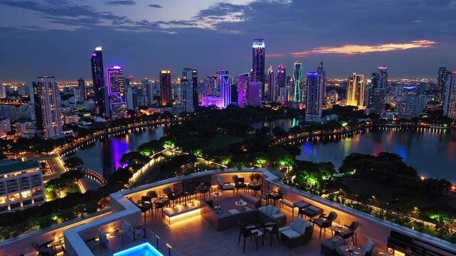 A breathtaking 4K ultra-HD aerial stock video capturing a stunning panoramic view of Lumpini Park and the Bangkok city skyline from a luxurious rooftop bar atop a high-end hotel. 