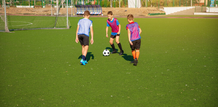 The photo shows three young soccer players in motion on a green artificial turf field, actively playing with a soccer ball.They wear colorful jerseys,shorts,and cleats, engaging in a practice session - Powered by Adobe