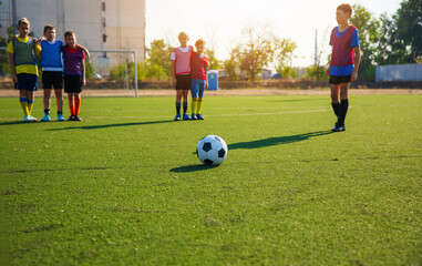 Obraz premium The photo shows a group of young soccer players in colorful jerseys standing in the background, while a soccer ball is placed in the foreground on a green turf field. The players are ready for action.