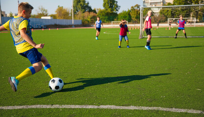 The photo shows young soccer player in yellow and blue uniform kicking a soccer ball on a green turf field.Other players in the background are actively engaged in the game. It is bright, dynamic scene