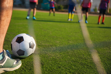 Obraz premium A close-up view of a soccer ball on a green field, with a player’s foot preparing to kick it. In the background, other players in colorful jerseys are seen practicing, with a clear blue sky.